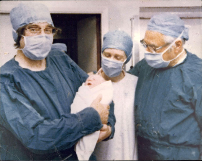 historical photo of three adults in hospital scrubs, caps and masks; one of them holds a baby