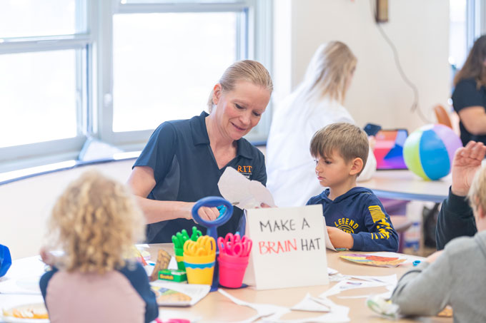 Rain Bosworth interacting with a child sitting next to her. Bosworth's blonde hair is pulled back into a ponytail, and she's wearing a short-sleeve navy shirt that displays the RIT logo in orange. The child. who has short blonde hair and wearing a navy hoodie, is watching Bosworth cutting a piece of paper with a pair of scissors. They are both sitting at a round table, which has a sign that reads, "Make a brain hat."
