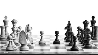 A black and white photograph from a low angle of a chess board with pieces set up for play. White chess pieces are on the left side of the board, while black pieces are on the right. In the center, a white pawn faces a black pawn.