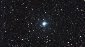 The exploding star V572 Velorum appears like a blue glowing orb with four rays extending out in a cross shape. It sits amid much dimmer blue and yellow stars and an inky night sky.