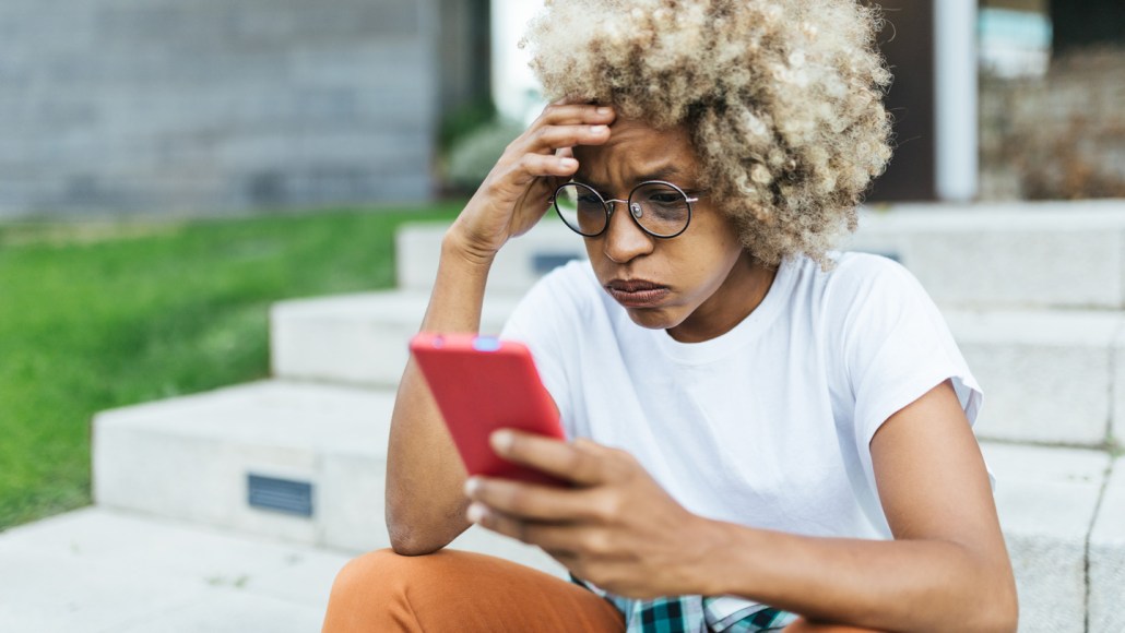 A young Black woman with light-colored hair sitting on stairs looks at her phone. She has a furrowed brow and her lips are pursed as if in worry or confusion. Her right hand is on her brow and her left holds a phone.