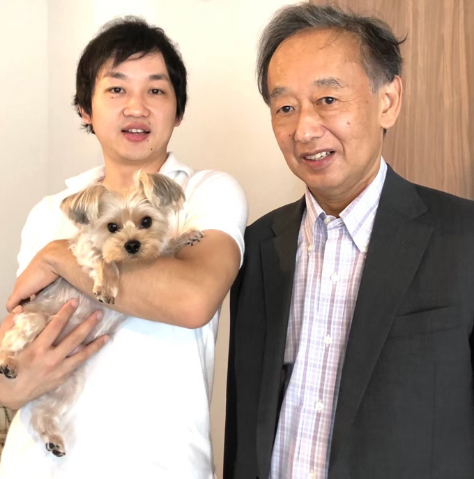 A photo of a young man holding a dog while standing to the left of an older man